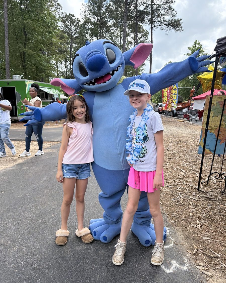 Two smiling girls posing with a big blue costumed character at the festival